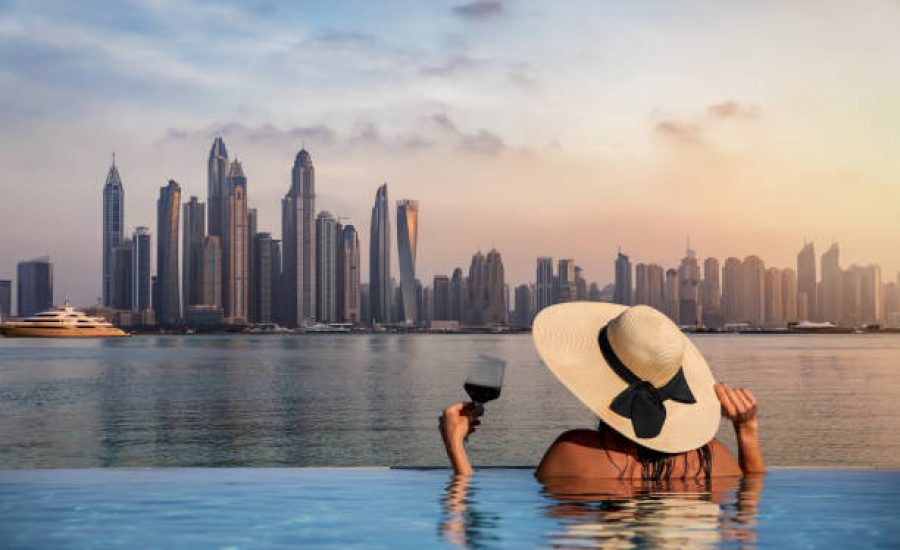 A elegant woman with a hat and a drink in her hand stands at the edge of a infinity pool and enjoys the view to the skyline of the Dubai Marina during sunset, UAE